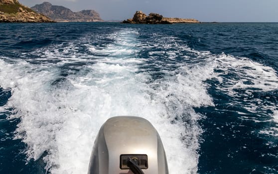 Stern wave of a motor boat with the motor in foreground and the rocky coastline on the eastside of Greek island Rhodes in the background on a sunny day in spring