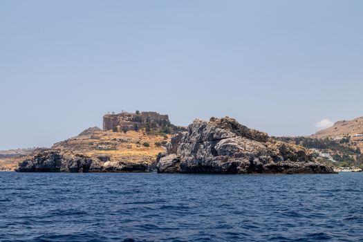 View from a motor boat on the mediterranean sea at the rocky coastline and the acropolis of Lindos near Stegna on the eastside of Greek island Rhodes on a sunny day in spring