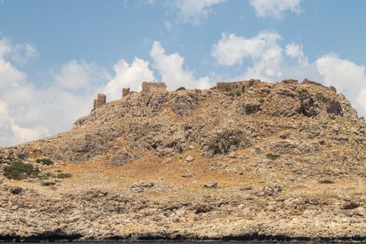 View from a motor boat on the mediterranean sea at the rocky coastline near Lindos  on the eastside of Greek island Rhodes with ruins of a castle on the top of a mountain