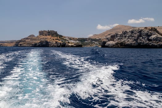 View from a motor boat on the mediterranean sea at the rocky coastline and the acropolis of Lindos near Stegna on the eastside of Greek island Rhodes on a sunny day in spring