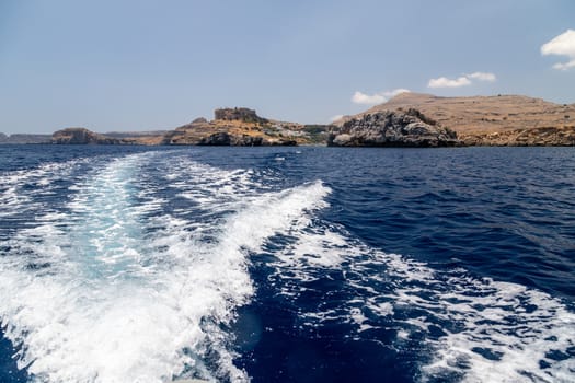 View from a motor boat on the mediterranean sea at the rocky coastline and the acropolis of Lindos near Stegna on the eastside of Greek island Rhodes on a sunny day in spring