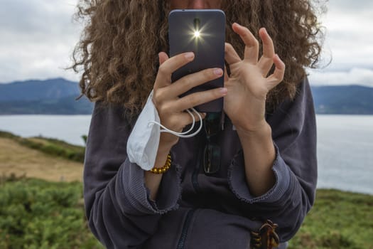 A young woman, grasping a protective face mask with her fingers, takes, at the same time, a photograph with a smart mobile phone, Spain.