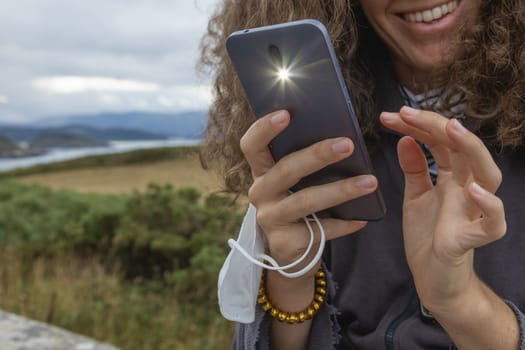 A young woman, grasping a protective face mask with her fingers, takes, at the same time, a photograph with a smart mobile phone, Spain.