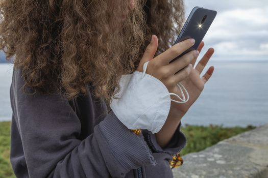 A young woman, grasping a protective face mask with her fingers, holds at the same time, with her hands, a smart mobile phone, Spain.