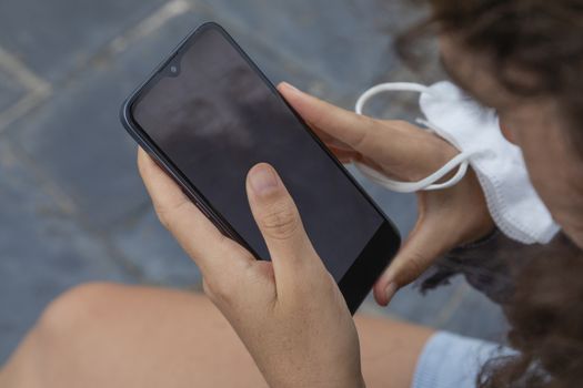 A young woman, grasping a protective face mask with her fingers, holds at the same time, with her hands, a smart mobile phone, Spain.