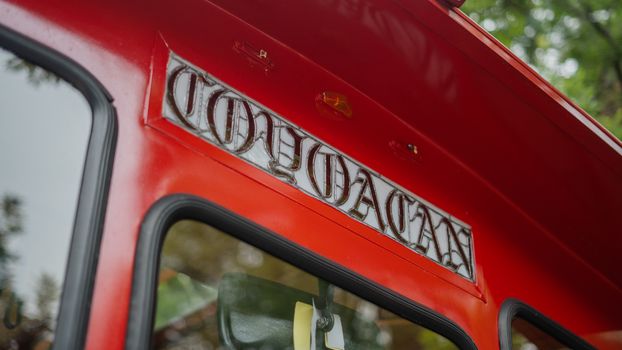 Picture of a Coyoacan Sign on a red and yellow trolley car driving on a street, with trees in the background. The text 'Coyoaca' is the name of the neighborhood in both Spanish and English