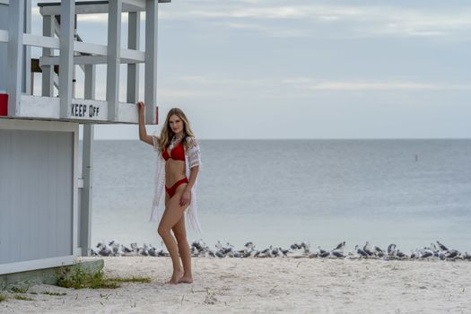 A beautiful blonde bikini model enjoys the weather outdoors on the beach while posing near a lifeguard station