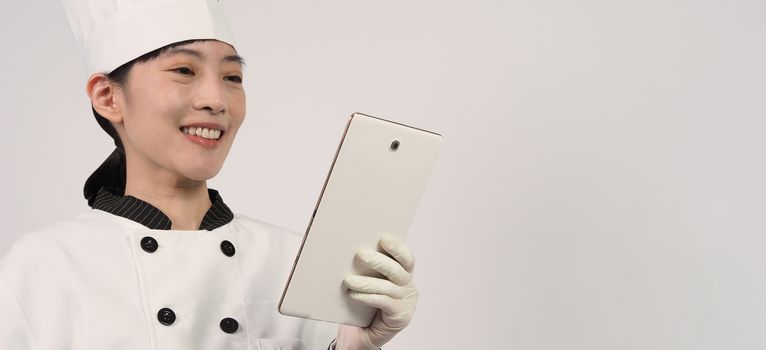 Asian woman chef holding smartphone or digital tablet and received food order from online shop or merchant application. she smiling in chef uniform and standing in studio with white color background.
