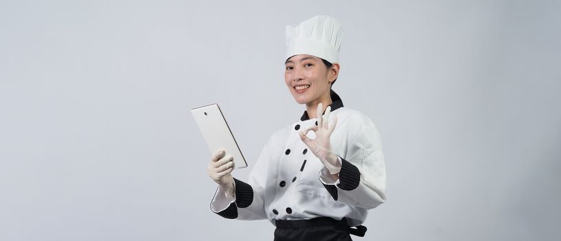 Asian woman chef holding smartphone or digital tablet and received food order from online shop or merchant application. she smiling in chef uniform and standing in studio with white color background.