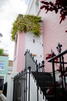 Portrait style picture of a pink building with railed stairs and plants hanging over its white door