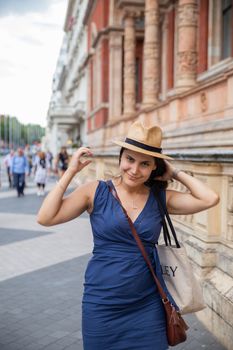 Portrait style picture of a smiling woman in a blue dress looking at the camera while she grabs her hair, and walks next to a building with classic architecture -South Kensington, London, UK-
