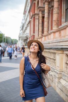 Portrait style picture of a smiling woman in a blue dress and a khaki hat happily looking up at the cloudy sky, with a building with classic architecture next to her -South Kensington, London, UK-