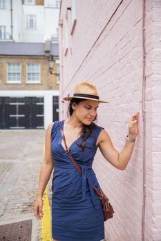 Portrait style picture of a woman in a blue dress and a khaki hat that has a reflexive look on her face when she touches a pink brick wall -South Kensington, London, UK-