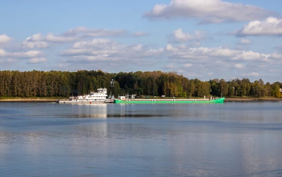 The boat pushes the barge ahead of it along the river along the shore.