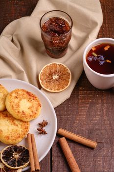 Cottage cheese fritters with hot black aromatic tea with fig jam, Christmas breakfast mood with anise, cinnamon and dried citrus on wooden background, angle view