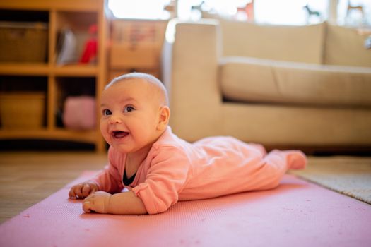 Picture of a smiling baby in a pink sleepsuit lying face down on a pink rug with a couch and a window as background