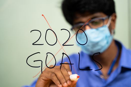 Young man in medical mask scratching out 2020 goals due to coronavirus or covid-19 pandemic - concept of failed 2020 goals
