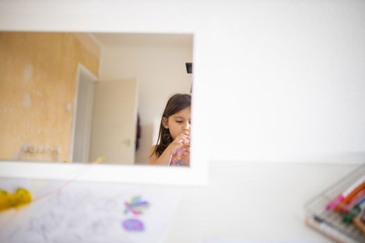 Little brunette girl and a white open door reflected on a mirror across a white table with a drawing of colorful leaves