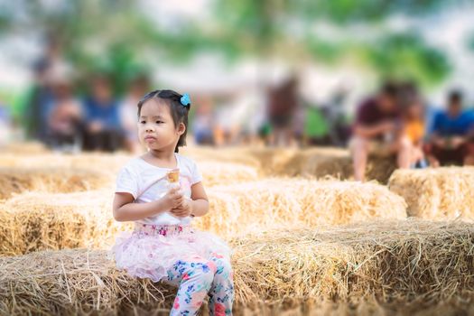 Asian little girl sitting on straw seat and eating an ice cream outdoors with people blur background