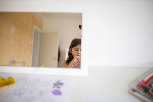 Little brunette girl and a white open door reflected on a mirror across a white table with a drawing of colorful leaves