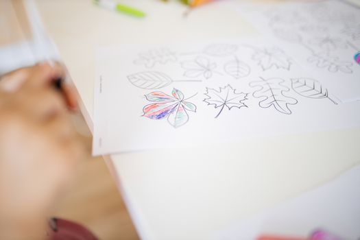 Paper sheet with coloring images of different types of leaves on a white table alongside the blurry hands of a little girl