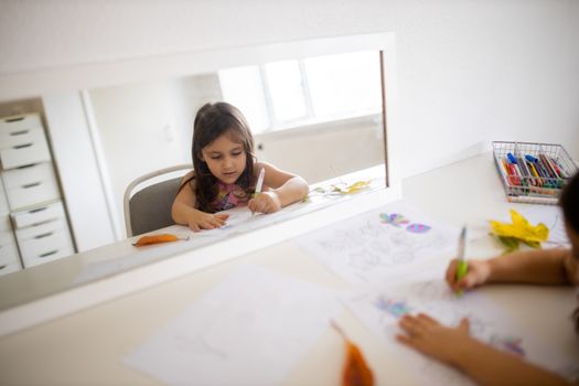 Landscape picture of a mirror across a white table reflecting a little girl in a pink dress coloring different types of leaves on a paper sheet, with real leaves aside