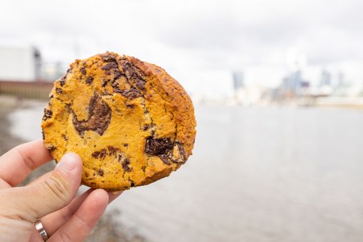 Landscape View of a Hand Holding a Chocolate Chips Cookie with a Blurry River Thames and CIty as Background