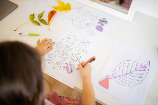 Over the head of a little brunette girl in a pink dress sitting in front of a mirror and coloring leaves