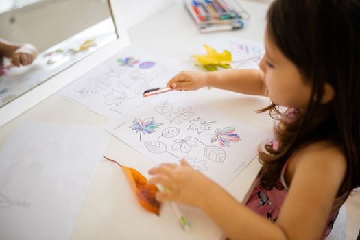 Landscape picture of a little brunette girl in a pink dress in front of a mirror coloring different types of leaves on a paper sheet, with real leaves aside