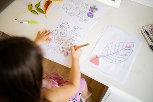 Over the head of a little brunette girl in a pink dress sitting in front of a mirror and coloring leaves images on paper