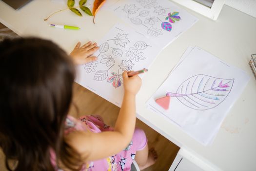 Over the head of a little brunette girl in a pink dress sitting in front of a mirror and coloring leaves
