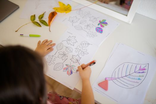 Over the head of a little brunette girl in a pink dress sitting in front of a mirror and coloring leaves