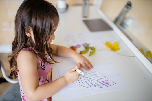 Little brunette girl in a pink dress sitting in front of a mirror and coloring a big leaf image on a paper sheet