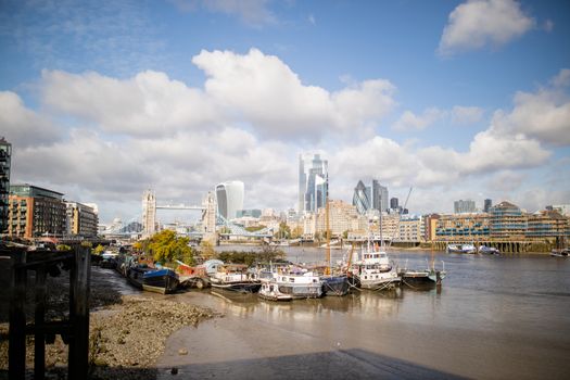 Landscape View of Docked Boats on the River Thames alongside a sandy and rocky ground, and with the Tower Bridge and a Cityscape as Background