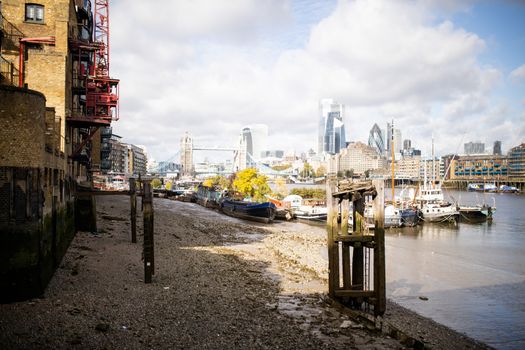 Landscape View of Docked Boats on the River Thames alongside a sandy and rocky ground, and with the Tower Bridge and a Cityscape as Background