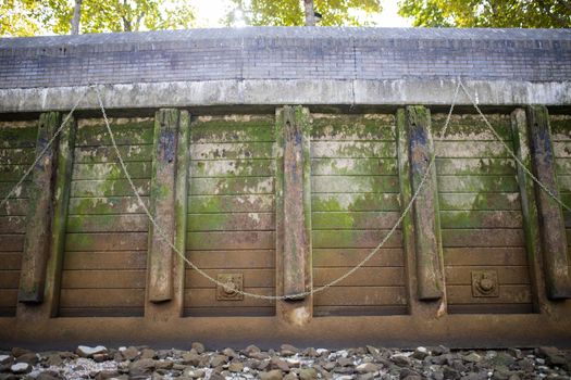 Landscape View of a brown Concrete Wall Partially Covered by Moss, with chains hanging from it, and a ground covered by gray rocks at the bottom