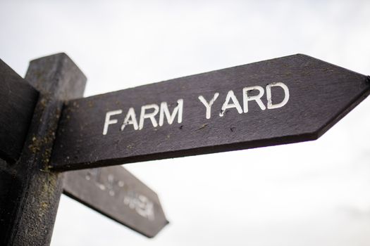 Wooden pole with indications pointing out the way to the farm yard and to other places, and with a cloudy sky as background