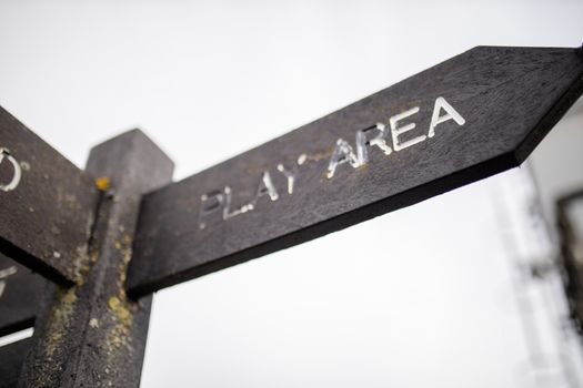Up close view of a wooden pole with indications pointing out the way to the play area and to other places, and with a cloudy sky as background