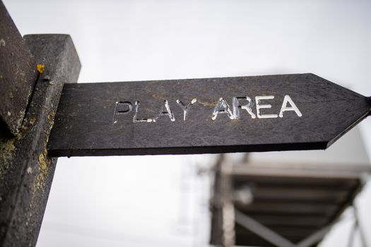Up close view of a wooden pole with indications pointing out the way to the play area, with a cloudy sky and a blurry structure as background