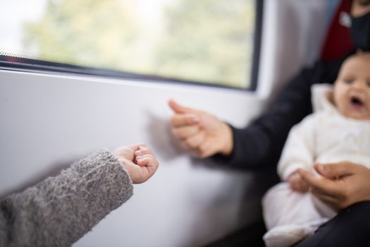 Picture of the arm of a little girl in a gray sweater receiving pistachios from her mother who sits next to a window in a bus, and holds her yawning baby daughter in her arms