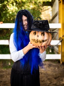 A girl dressed as a witch with blue hair is holding a pumpkin with a painted face and a witch's cap