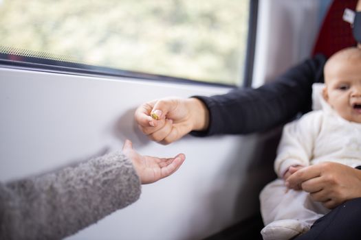 Picture of the arm of a little girl in a gray sweater receiving pistachios from her mother who sits next to a window in a bus, and holds her baby daughter in her arms