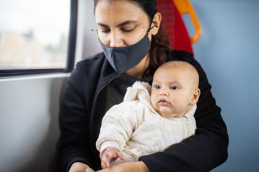 Woman wearing a face mask and looking down while sits next to a window in a bus and holds her baby