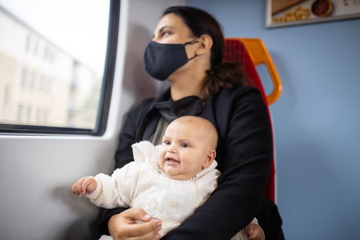 Woman wearing a face mask and black clothes, looking through the window of a bus as she holds her smiling baby