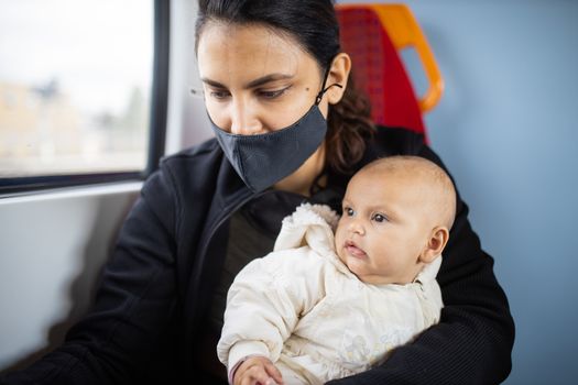 Woman wearing a face mask sitting next to a window in a bus and holding her distracted-looking baby