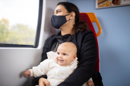 Woman wearing a face mask and black clothes and looking through the window of a bus as she holds her smiling baby