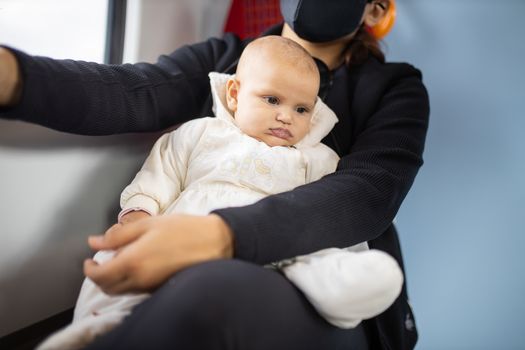 Distracted-looking baby in white clothing resting on the legs of her mother who wears a face mask and sits next to a window