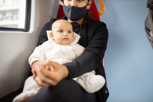 Baby in white clothing looking into the distance and resting on the legs of her mother who wears a face mask