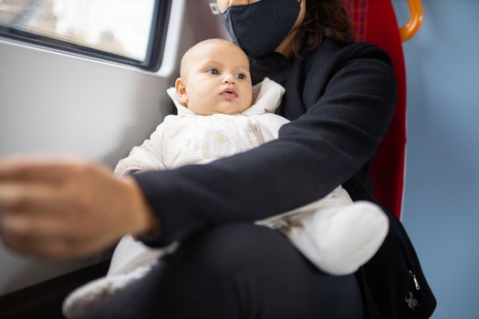 Distracted-looking baby in white clothing resting on the legs of her mother who wears a face mask and sits next to a window