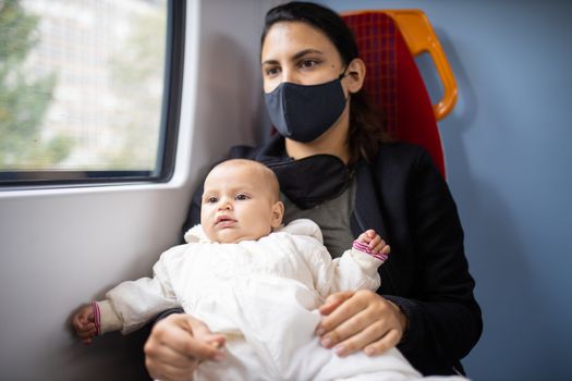 Woman wearing a black face mask sitting next to a window in a bus and holding her distracted-looking baby daughter